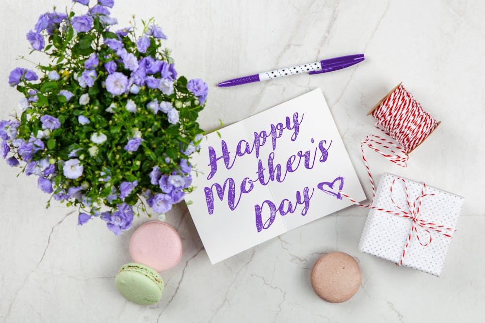 A flat lay of a "Happy Mother’s Day" card with purple flowers, macarons, a wrapped gift, and a pen on a marble surface.