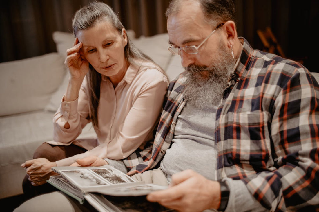 An elderly man and woman sitting closely on a couch, thoughtfully looking through a black-and-white photo album together.
