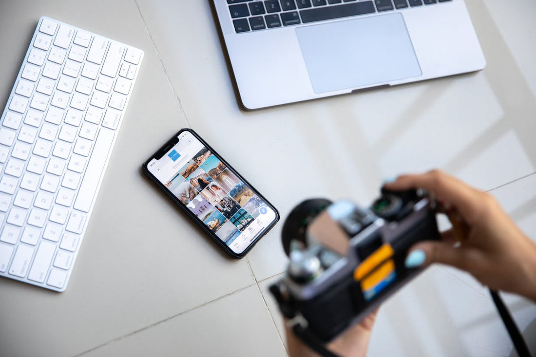 Person holding a camera while organizing digital photos displayed on a smartphone, with a laptop and keyboard nearby.