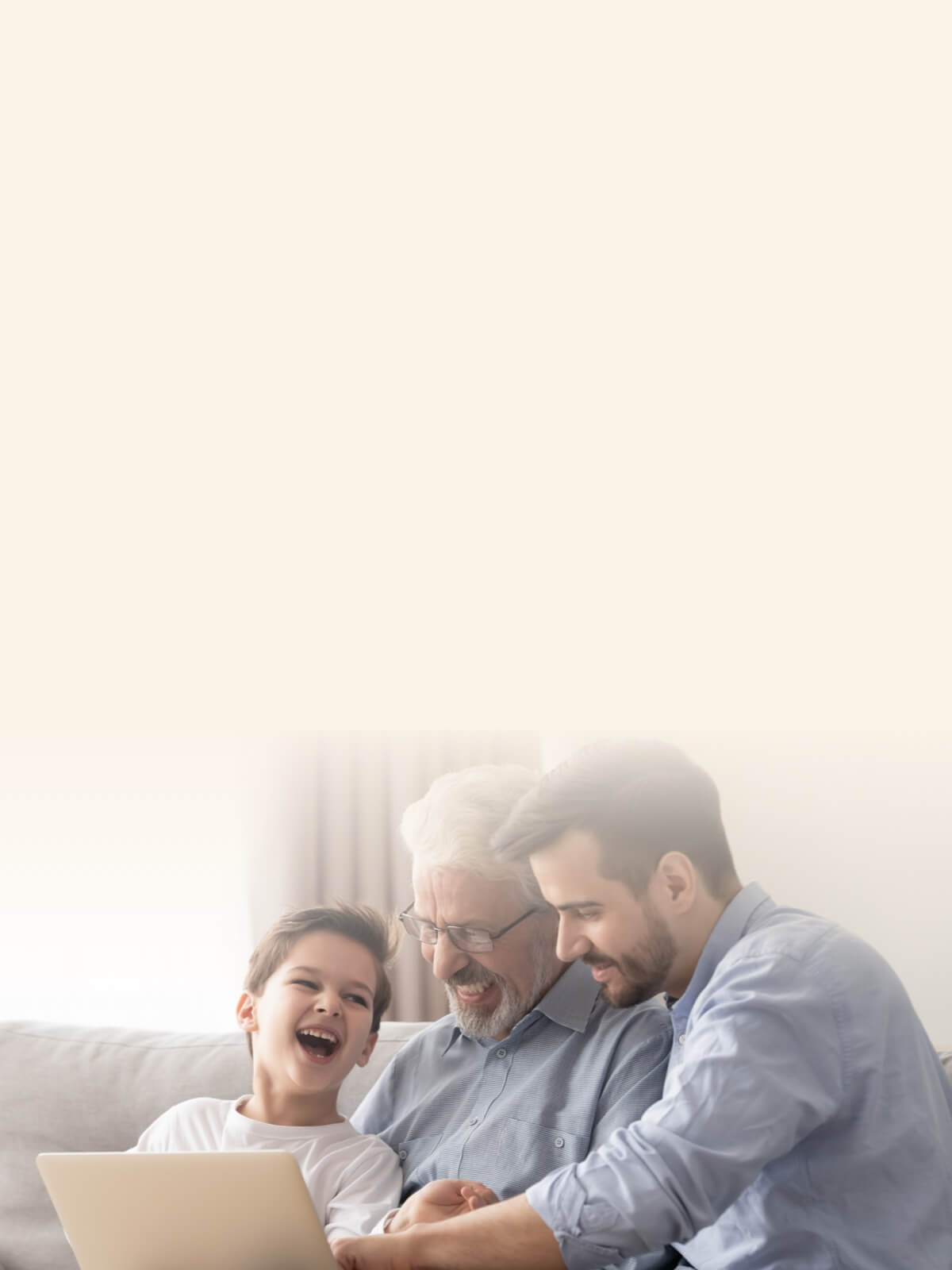 A grandfather, father, and son sitting together on a couch, smiling and looking at a laptop.
