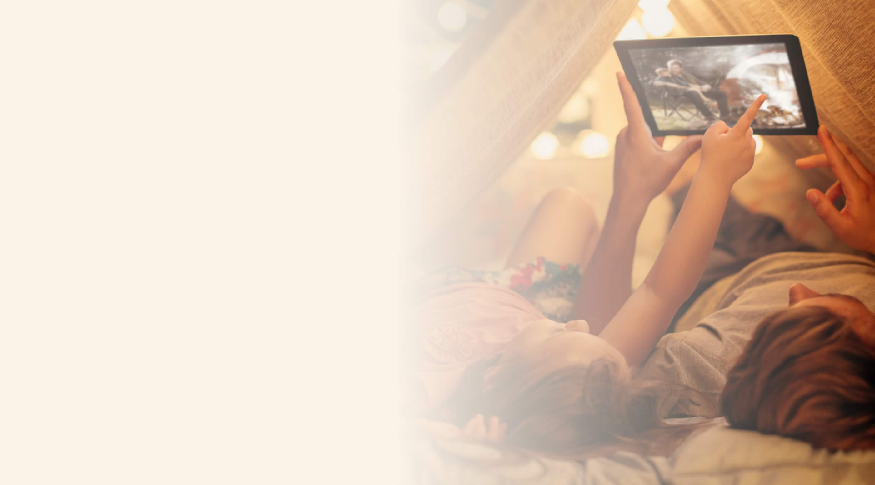 A parent and child lying inside a blanket fort, pointing at a tablet screen showing a person by a campfire.