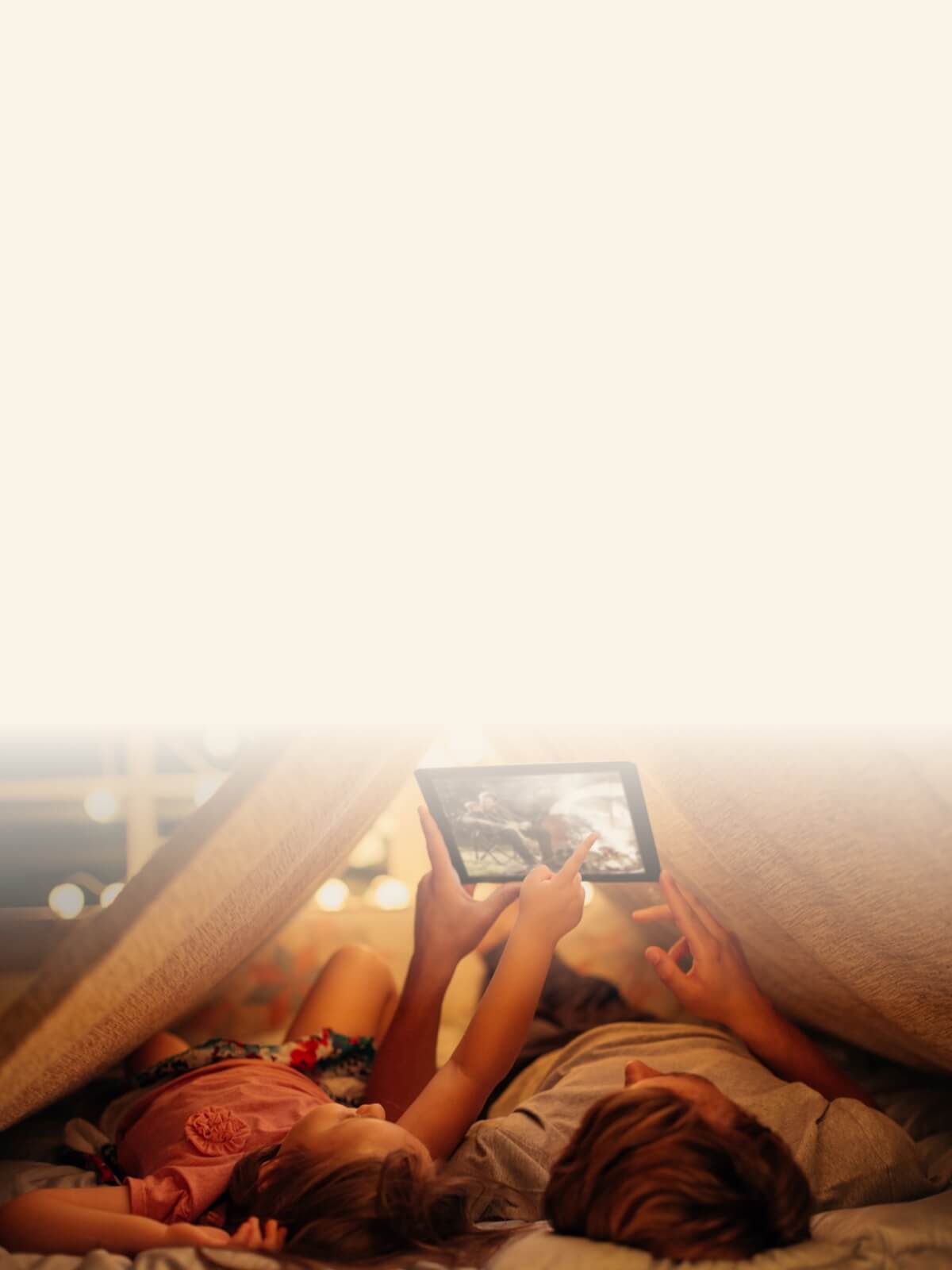 A parent and child lying inside a blanket fort, pointing at a tablet screen showing a person by a campfire