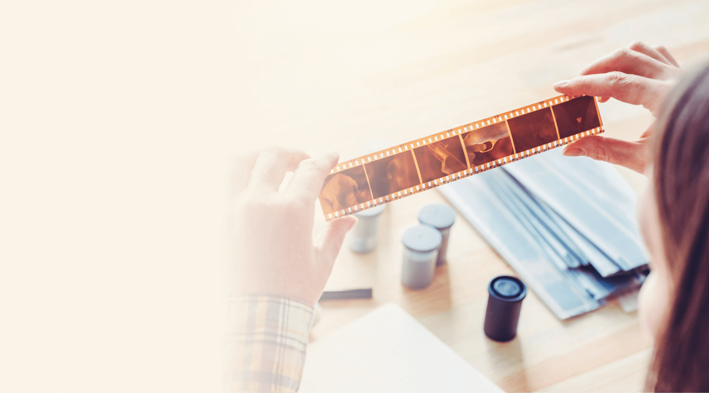A person holding a strip of film negatives up to the light, with film rolls and protective sleeves on the table.